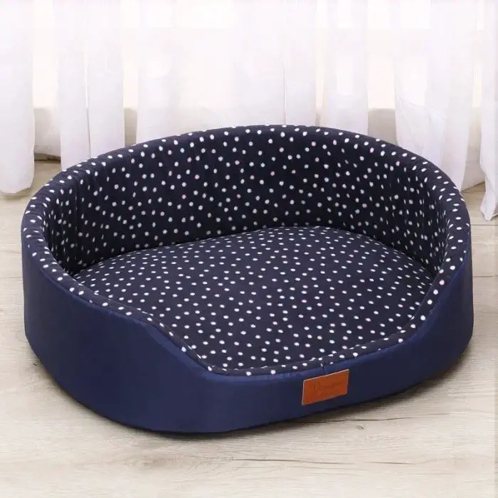 A cozy, round, dot-patterned pet bed in navy blue with white polka dots sits on a wooden floor in front of white curtains called 10233-4a6a87.jpg.