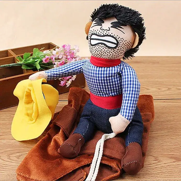 A fabric cowboy doll with a grimacing face sits on a wooden surface, holding a yellow hat. The 10673-4ca322.jpg wears a blue checkered shirt, red belt, jeans, and boots that complete his costume.