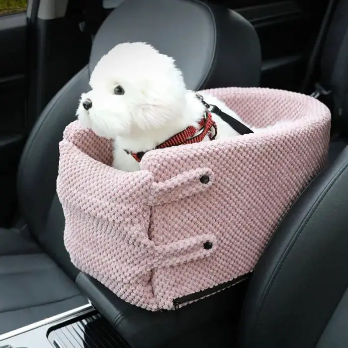 A small, fluffy white dog sits in a pink, textured 10946-79c9a9.jpg secured with black buckles, positioned on the passenger seat of a car.