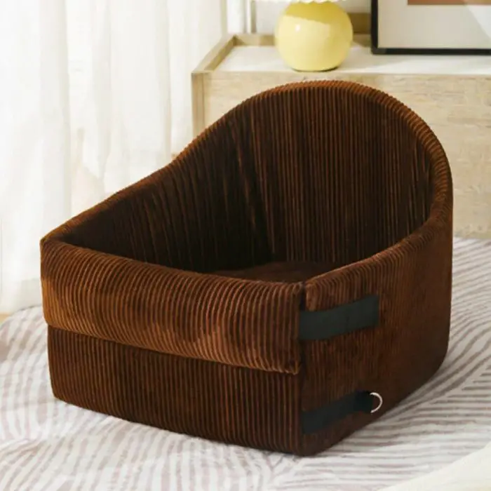 A brown, ribbed fabric pet bed resembling 11114-4bab88.jpg with an elevated backrest sits on a striped surface next to a wood table and a yellow vase.