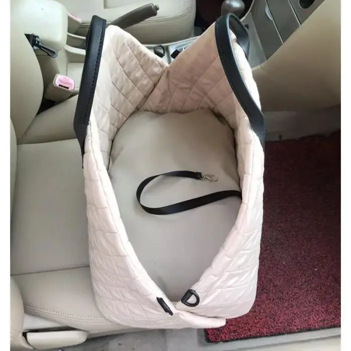 An empty, beige quilted 11115-c3dddb.jpg pet car seat with black edges and a built-in safety strap is placed on the passenger seat of a car. The light-colored interior features a red floor mat that's easily visible.