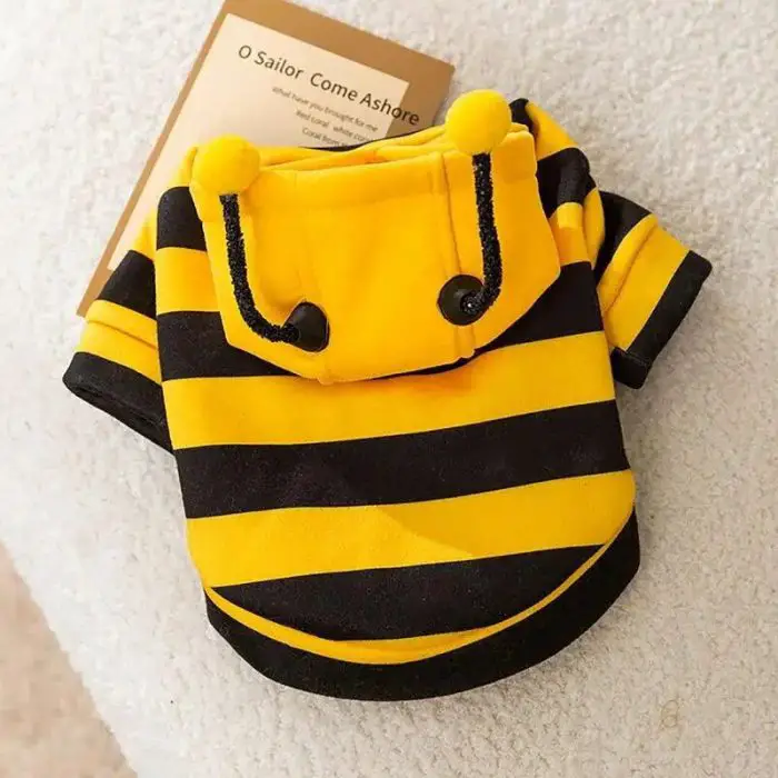 A small, yellow and black striped pet hoodie with a hood and playful pom-poms, resembling a bee costume hoodie, placed on a white surface. A brown tag with text is attached to 11487-f649a4.jpg.