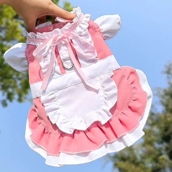 A pink and white frilly costume for pets, revealed as product 11844-e32498.jpg, is held up against a backdrop of trees. The dress showcases ruffles, a ribbon bow, and an apron-like design.