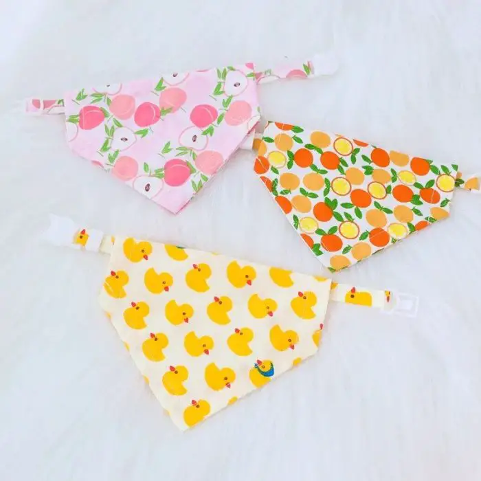 Three baby bibs are displayed on a white surface: one with a pink fruit pattern, one with an orange citrus pattern, and one with a yellow rubber duck pattern. The product, 11880-5a28de.jpg, offers these bandana bibs that add both style and functionality to your baby's wardrobe.