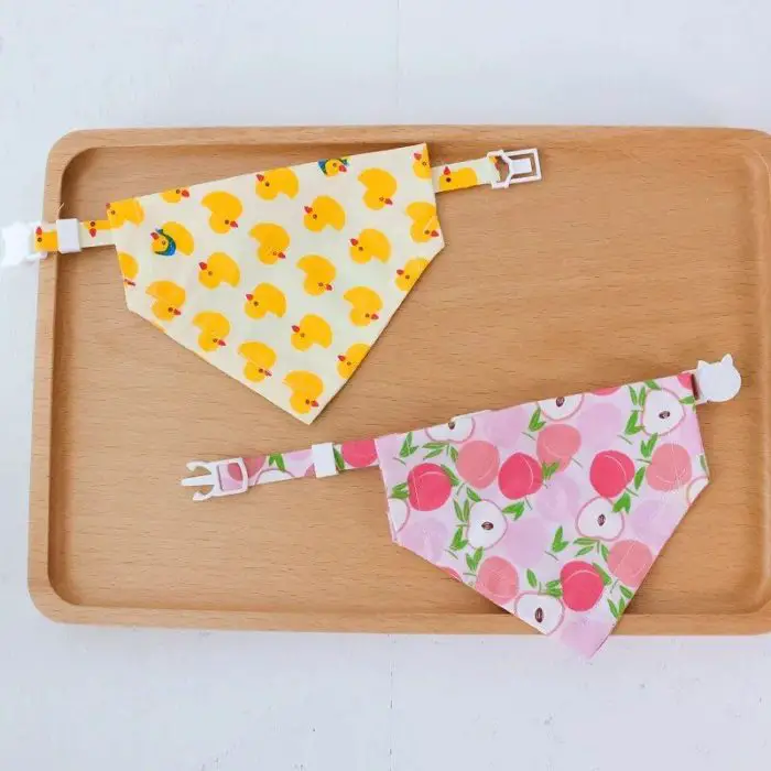 Two fabric bandanas with adjustable straps sit on a wooden tray. The top bandana displays a cheerful yellow duck pattern, while the bottom one features a charming pink and white peach design. Product Name: 11880-f70db5.jpg.