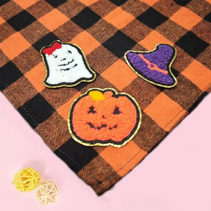 An orange and black plaid bandana adorned with three Halloween-themed patches (a ghost, a witch hat, and a pumpkin), complemented by two small beige woven balls, as depicted in product image 11881-049d34.jpg.