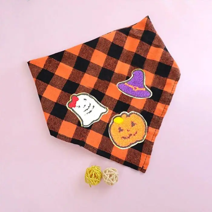 An orange and black checkered bandana, embellished with embroidered Halloween-themed patches depicting a ghost, a witch hat, and a pumpkin. Two decorative rattan balls are placed alongside on a light pink surface. Product: 11881-dbe3e9.jpg