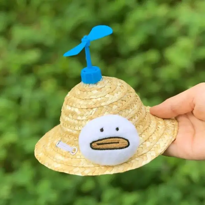 A hand holds the 12313-450335.jpg, a charming sun hat featuring a blue propeller on top and a small embroidered duck face on the front. The background showcases blurred green foliage.