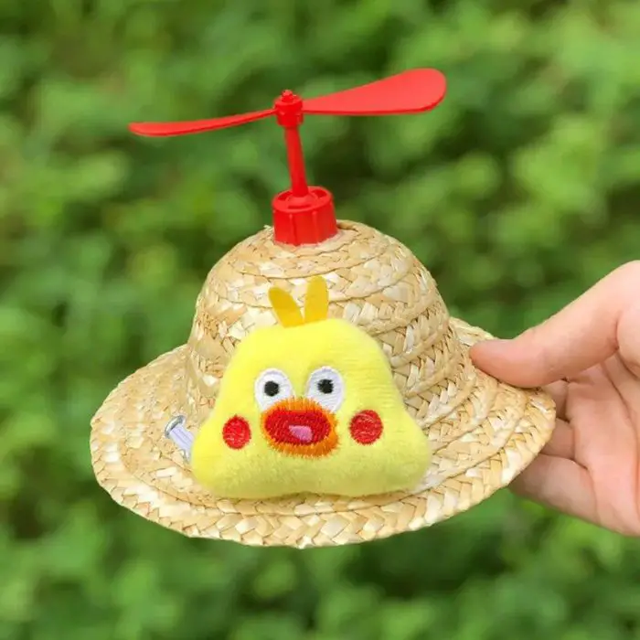 A hand holding "12313-6c56d1.jpg," a distinctive sun hat featuring a red propeller on top and a yellow plush toy with red cheeks and a surprised expression attached to the front. The background showcases vibrant green foliage.