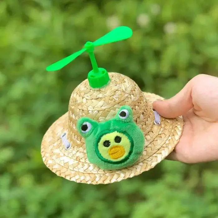 A hand holds the product 12313-6cc781.jpg, a straw sun hat adorned with a green plush frog face and a small green propeller on top, against a blurred green background.