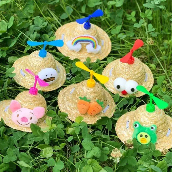 Six whimsical sun hats with propellers on top, each adorned with unique decorations including a rainbow, duck face, carrots, googly eyes, pig face, and a frog face, placed on green clover leaves (Product name: 12313-9e9945.jpg).