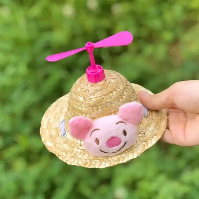 A hand displays the product "12313-b684b7.jpg," a charming small sun hat adorned with a plush pink pig face on the front and topped with a vibrant pink propeller, all set against a green, blurred background.