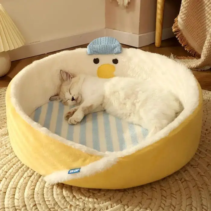 A white cat sleeps curled up in a round cat bed with a duck face design, featuring yellow and white colors. The cozy cat bed, depicted in image 12439-0e642f.jpg, rests on a woven rug in the inviting room.