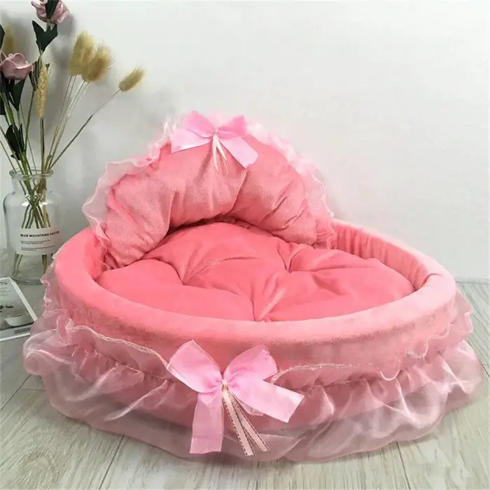 A pink, frilly dog bed with two bows, as seen in product 12440-99a152.jpg, is placed on a wooden floor. A vase with flowers is positioned beside it against a white wall.