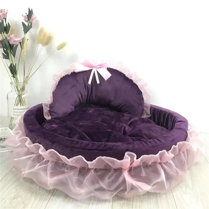 A plush purple dog bed (12440-aa4728) with a pink ruffled trim and a pink bow on the backrest, placed on a wooden floor beside a vase with flowers.