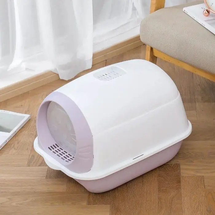 A covered white and light pink cat litter box (Product: 12586-33f776.jpg) sits on a wooden floor near a beige chair and a white curtain, creating a cozy nook.