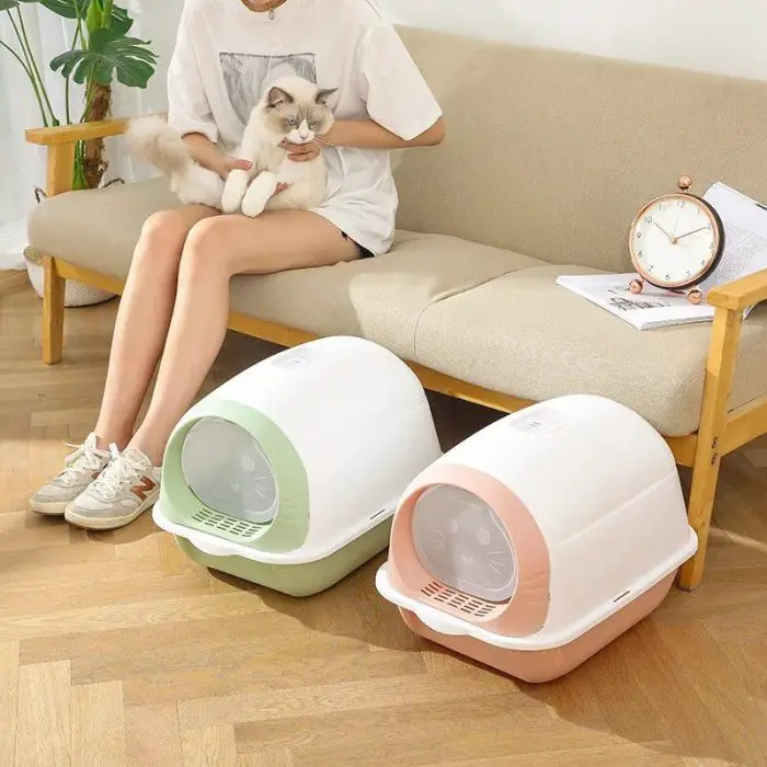 A person is sitting on a couch, holding a cat, with two dome-shaped cat litter boxes—one green and one pink—placed on the floor in front of them. An alarm clock and papers are situated on a cushion beside the person. The image is labeled as 12586-82d324.jpg.