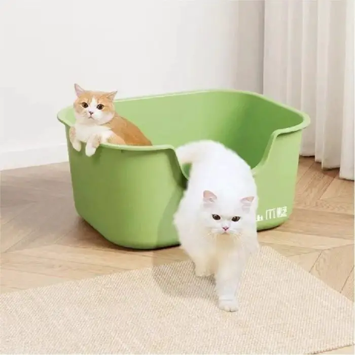 Two cats are near a green cat litter box labeled 12587-a8a988.jpg on a wooden floor. One orange and white cat is inside the container, while a white cat is walking out on a light-colored rug.