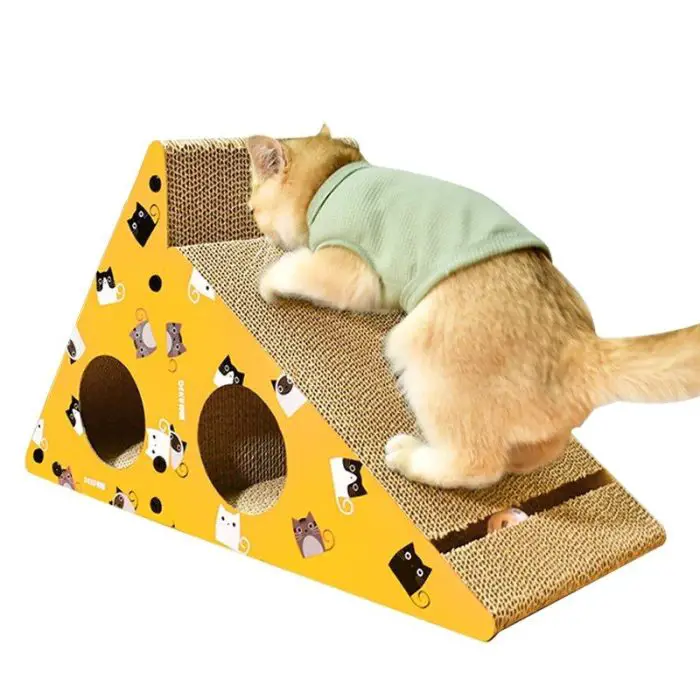 A cat wearing a green shirt climbs on product 12708-118242.jpg, a triangular cardboard cat scratcher adorned with cartoon cat designs and circular cutouts.