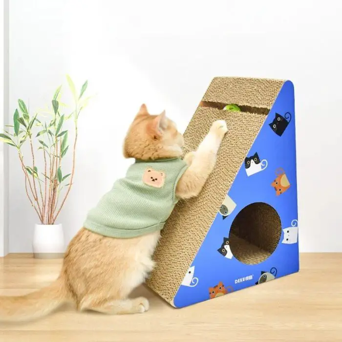 A cat wearing a green shirt interacts with the 12708-cba4e2.jpg cat scratcher, which features blue and brown colors. A small plant decorates the table behind the cat.