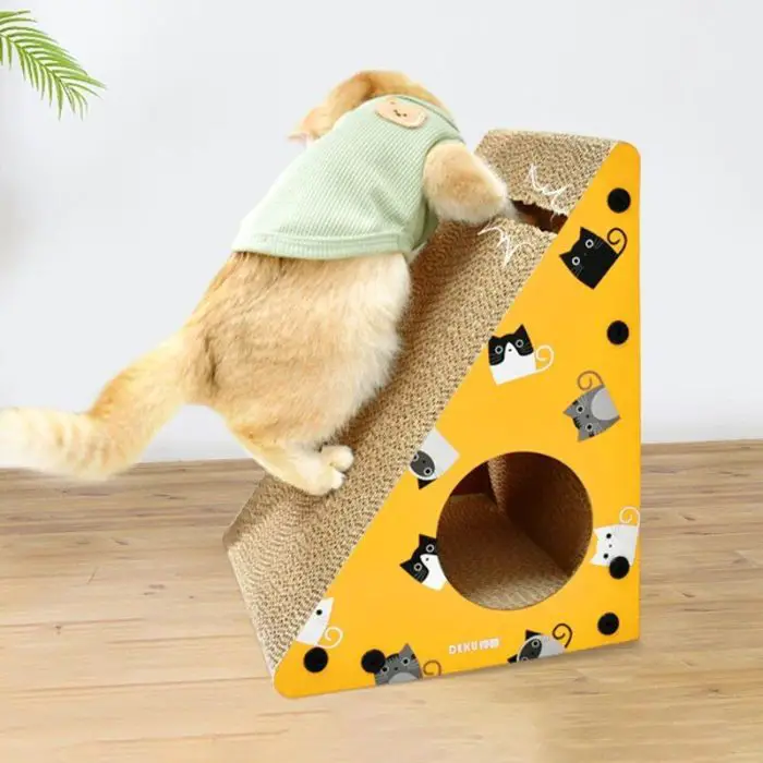 A cat wearing a green sweater climbs a yellow triangular scratcher adorned with cartoon cat faces and featuring a circular hole at the bottom (12708-dcab7f.jpg).