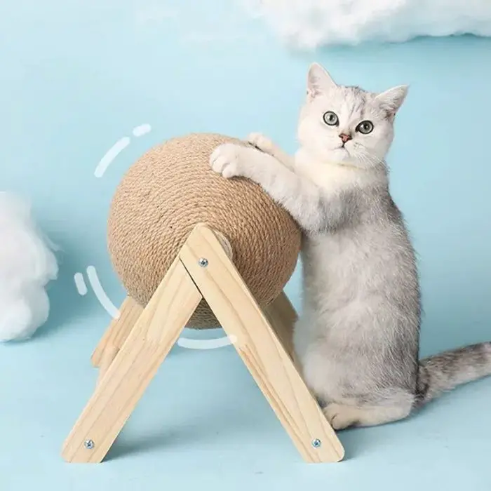 A cat stands on its hind legs with its front paws on a product from the 12730-abc7b1.jpg series, which is a spherical rope-covered post mounted on a wooden frame. The background features a light blue color with cloud-like decorations.