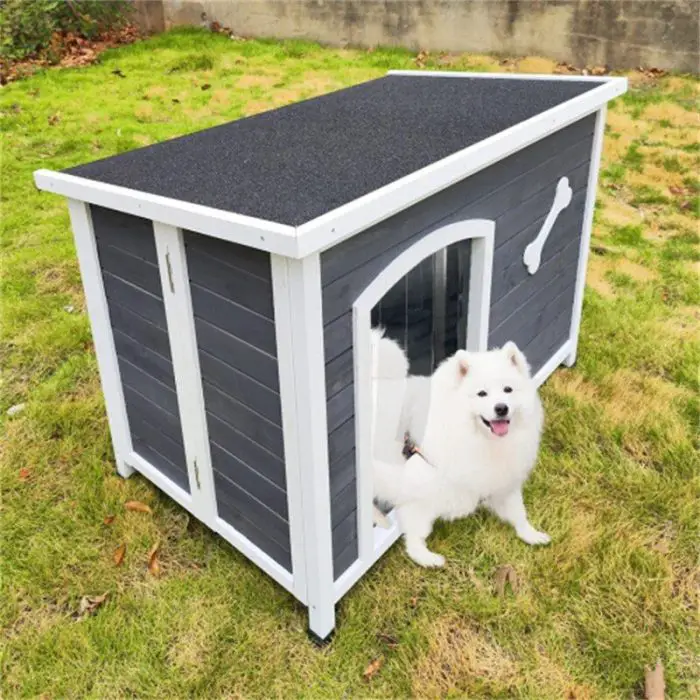 A white dog sits in front of the delightful 12744-38f999.jpg black and white dog house, which is situated on a lush grassy lawn and adorned with a bone decoration above its entrance.