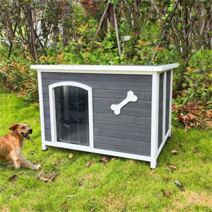 A gray and white dog house, complete with a bone decoration on the side, is placed on grassy ground with a dog sitting nearby. In the background, shrubs and trees create a perfect setting for the product 12744-744f67.jpg.