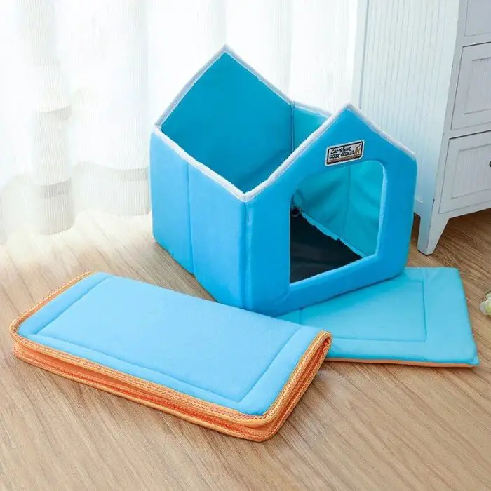 A blue, house-shaped dog bed (12784-39f356.jpg) with an open top is situated on a wooden floor. A detached orange-edged cushion rests in front of the pet bed. White curtains and a cabinet are in the background.