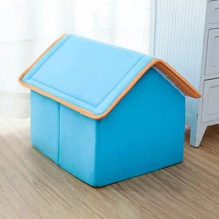 A product named 12784-797ae2.jpg, featuring a small blue fabric dog house with a triangular roof and orange trim, is placed on a wooden floor next to a white curtain and a cabinet.