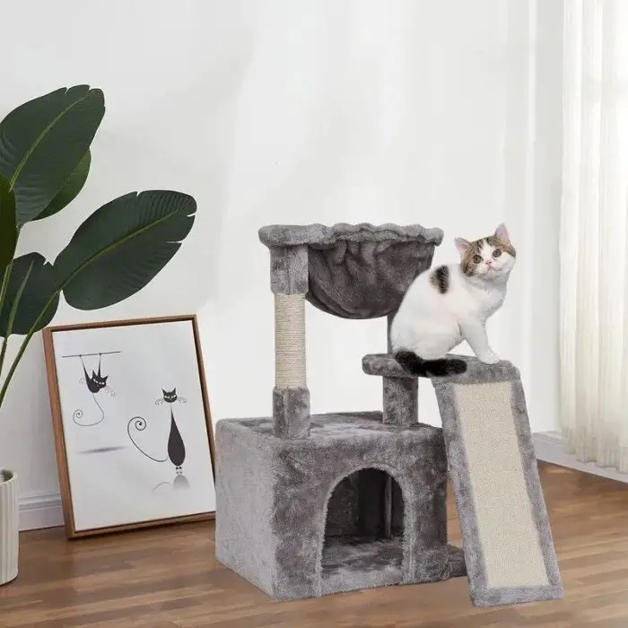 A white and gray cat relaxes on the 12807-850930.jpg Cat Activity Tower, which features multiple levels and a ramp, in a room with wooden flooring. In the background, there is a large plant and a framed picture adorned with cat illustrations.