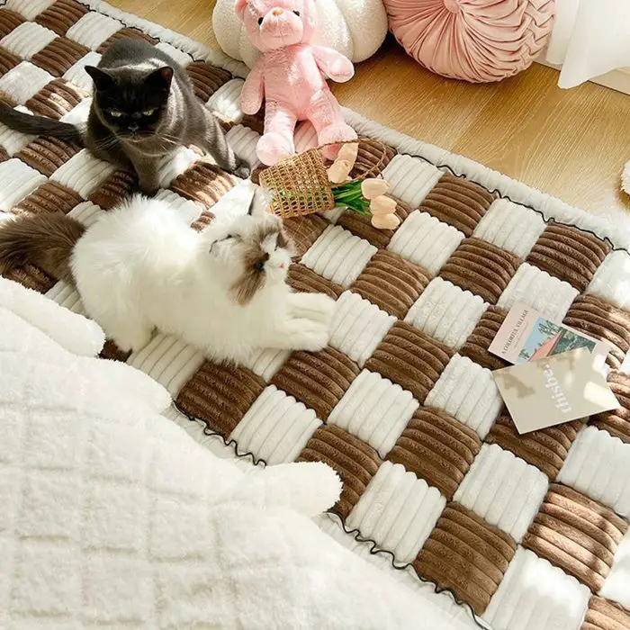 Two cats, one black and one white, relax on a 9523-077da9.jpg with a brown and white checkered pattern. The 9523-077da9.jpg features an anti-slip backing to keep it securely in place. Surrounding them are a pink stuffed toy, artificial flowers, and an open book.