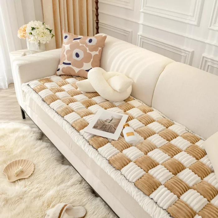 A luxurious cream and beige checkered throw with anti-slip backing covers a white sofa, adorned with a beige floral cushion and a cream pillow. Nearby are a magazine, 9523-9973e9.webp, white curtains, flowers in a vase, and a rug.