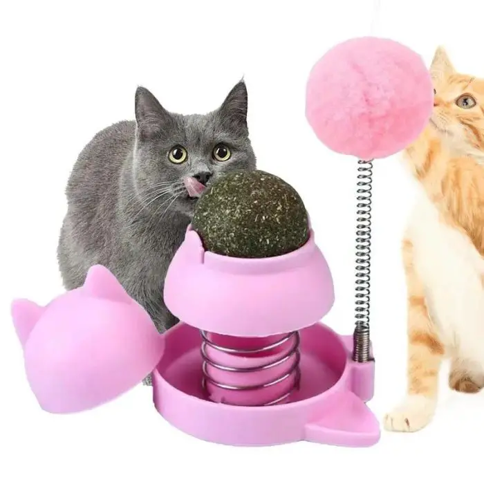 Two cats interact with 9783-57f7c2.jpg. One cat licks an adhesive catnip ball on a spring, while the other cat looks at a pink fluffy ball attached to the toy.