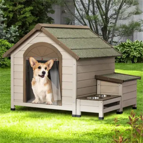 A dog, likely a Corgi, sits in a small, beige 9869-6968ff.jpg with a green roof. The cozy 9869-6968ff.jpg is outdoors in a yard with lush greenery. Next to the 9869-6968ff.jpg are attached food and water bowls.