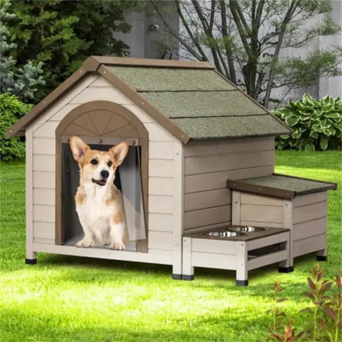 A dog, likely a Corgi, sits in a small, beige 9869-6968ff.jpg with a green roof. The cozy 9869-6968ff.jpg is outdoors in a yard with lush greenery. Next to the 9869-6968ff.jpg are attached food and water bowls.